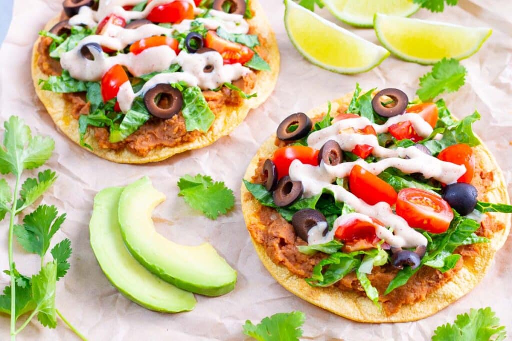 Two vegan tostadas on parchment paper with sliced avodado, lime wedges, and cilantro around them.