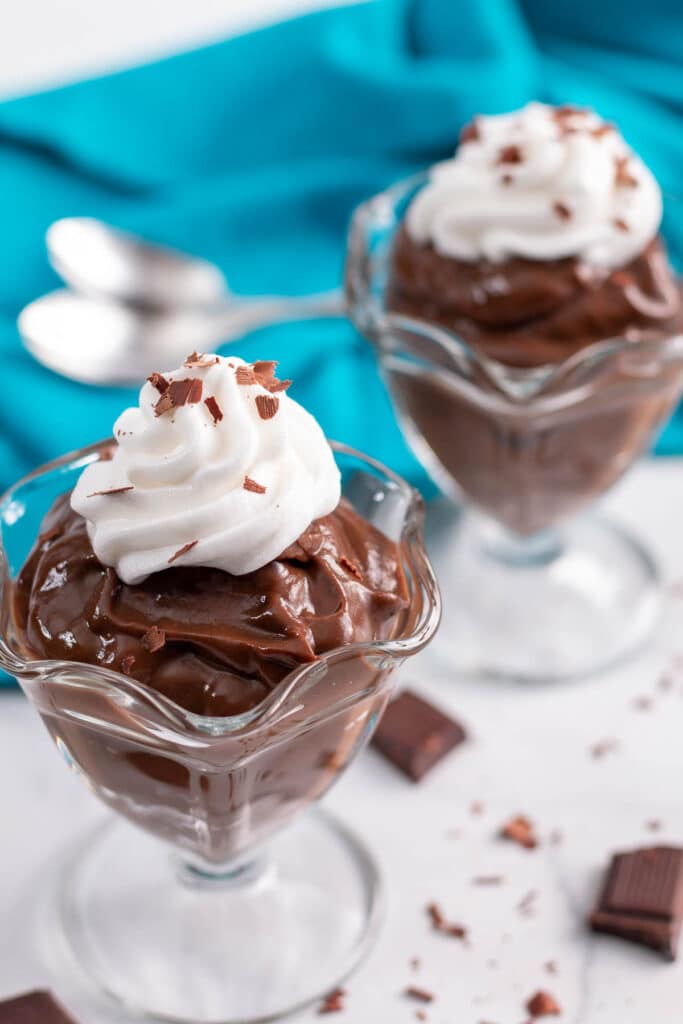 Two dessert cups of cocolate pudding with whipped cream and chocolate shavings surrounded by more chocolate, blue napkin, and spoons.