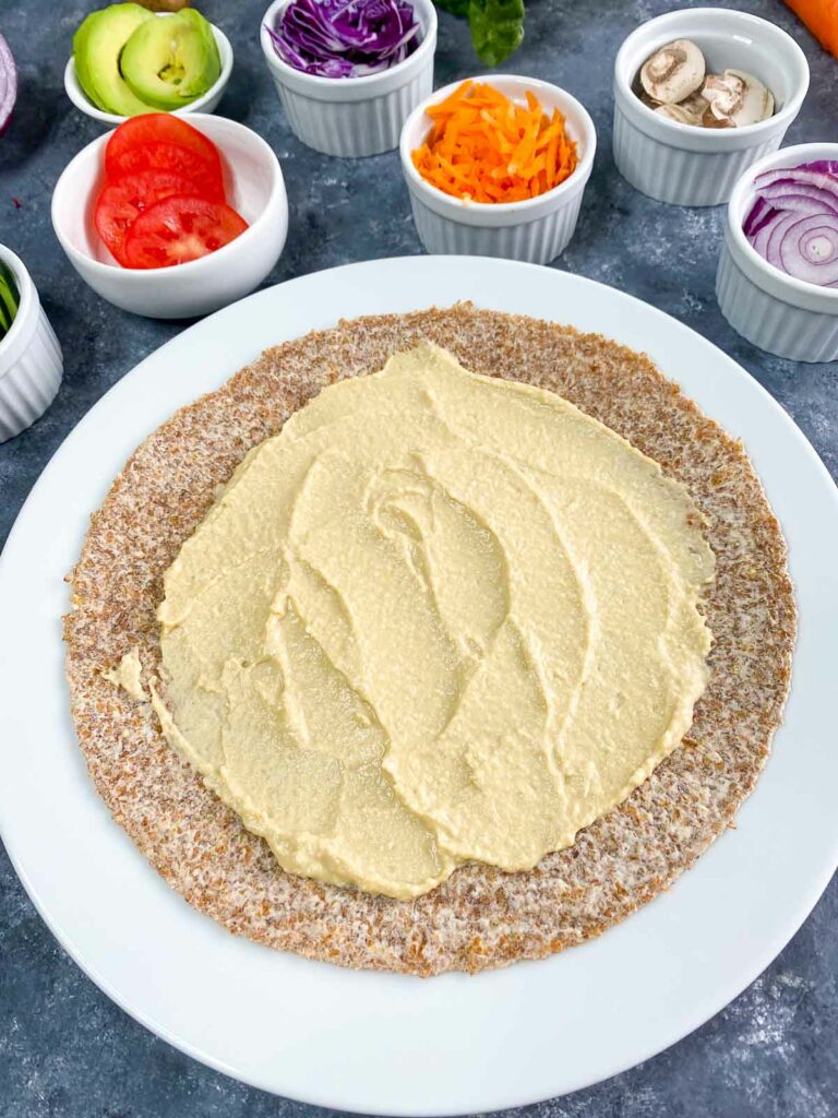 Hummus spread on tortilla with veggie wrap ingredients in the background.