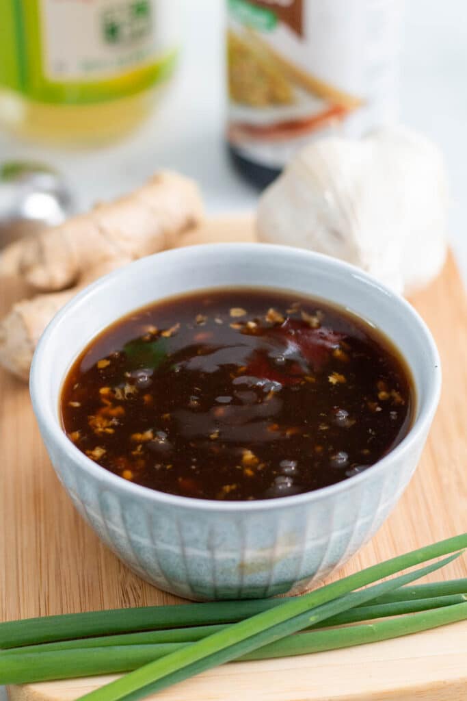 Bowl of teriyaki sauce on a wooden cutting board, surrounded by green onions, fresh garlic, and fresh ginger, with bottles of soy sauce and vinegar in the background.