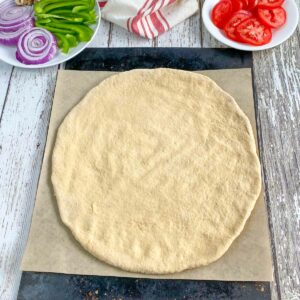 Pizza dough spread out on parchment lined pan with plates of pizza toppings in background.
