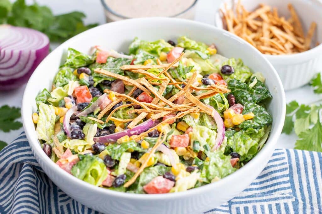 Bowl of salad on a blue and white striped napkin with salad ingredients in the background.