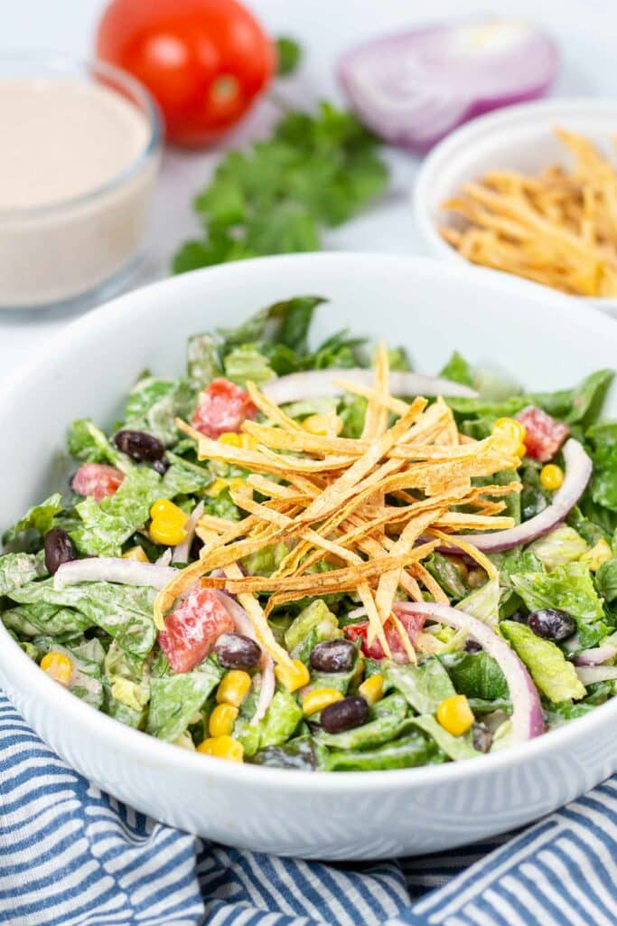 Bowl of salad topped with tortilla strips on a blue and white striped napkin with salad ingredients in the background.