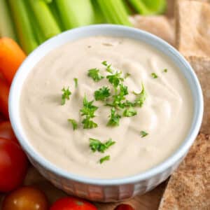 Close up image of bowl of tahini sauce garnished with parsley and surrounded by pita bread, tomatoes, carrots and celery.
