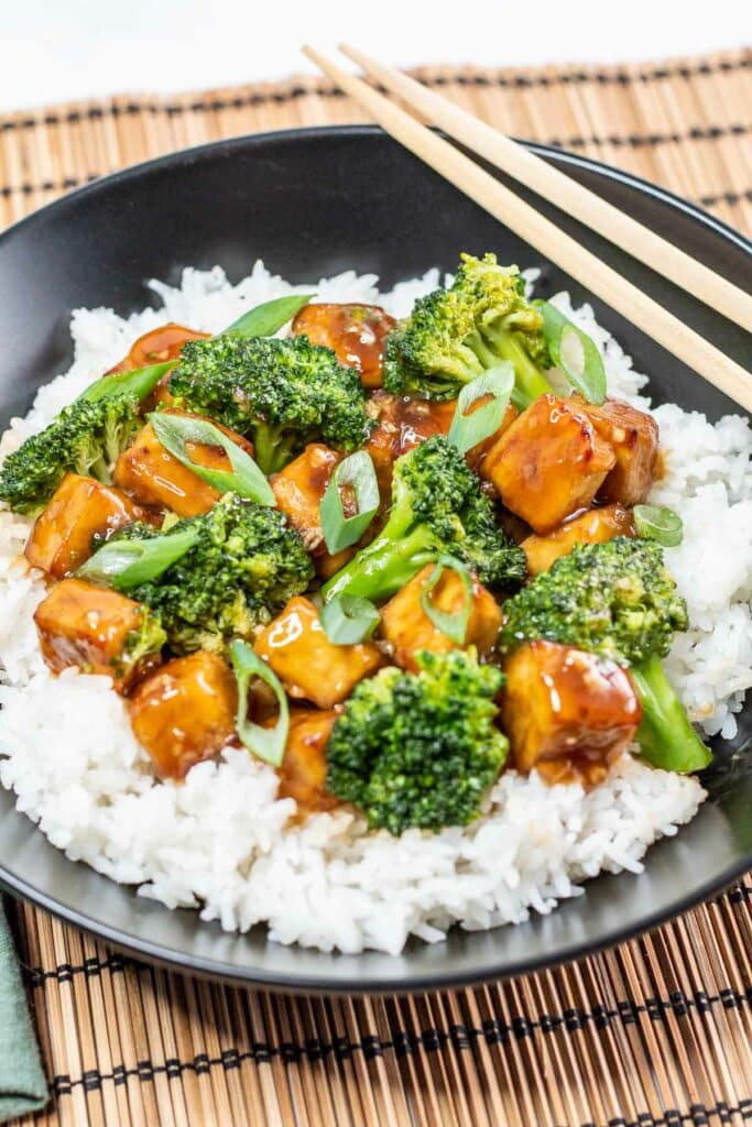 Bowl of teriyaki tofu and broccoli on rice with chopsticks resting on side of bowl.