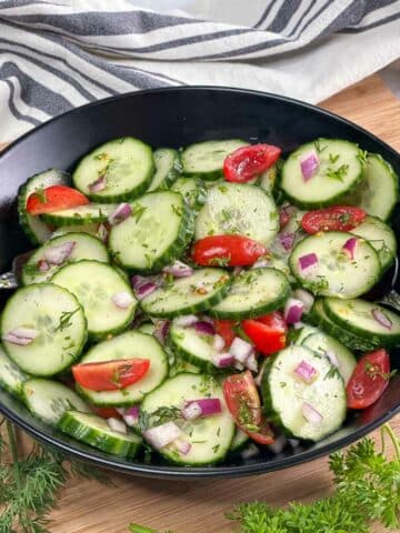 Cucumber salad in a black bowl with striped napkin and fresh herbs around it.