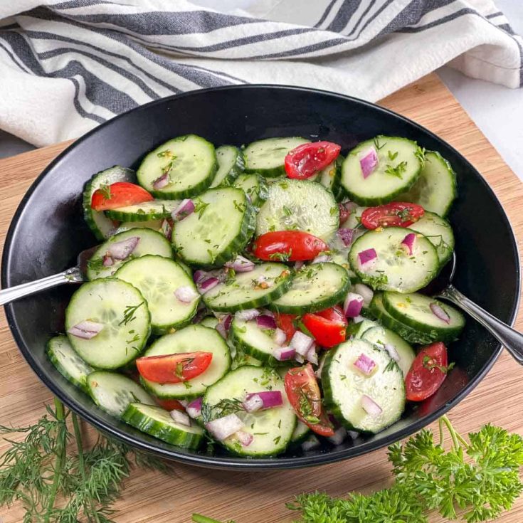 Cucumber salad in a black bowl with striped napkin and fresh herbs around it.