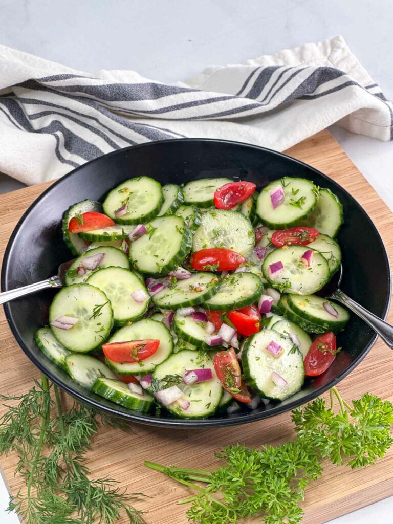 Cucumber salad in a black bowl surrounded by cloth napkin and fresh herbs.