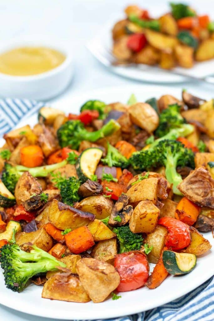Roasted vegetables on a white platter with serving plate of vegetables and bowl of sauce in the background.