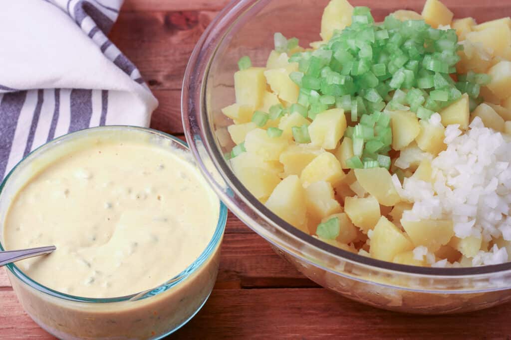 Large bowl of potatoes, celery, and onion with a small bowl of potato salad dressing next to it.