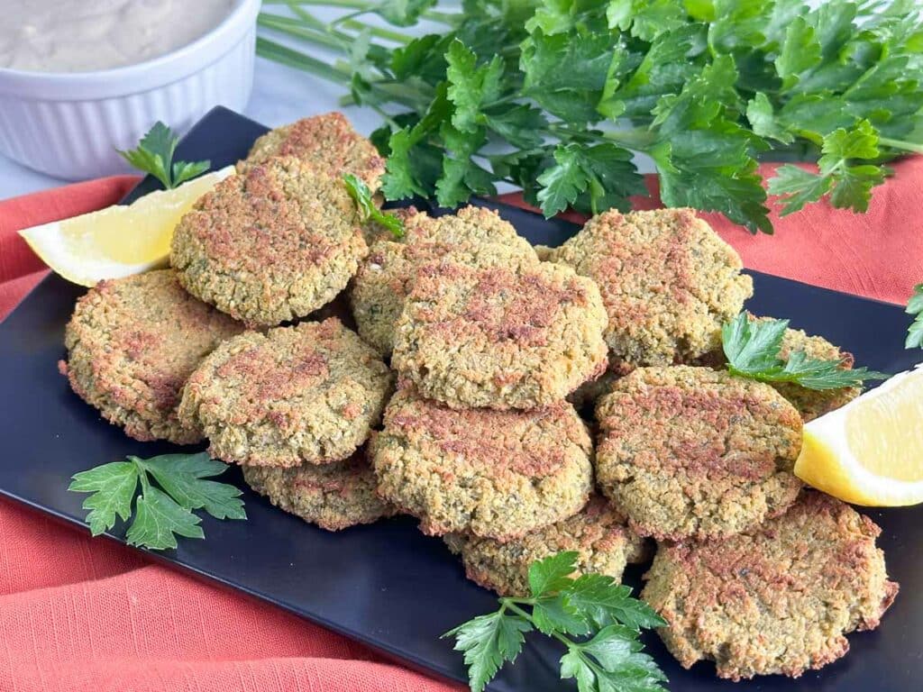 Falafel stacked on a black platter garnished with lemon wedges and parsley with bunch of pasley and bowl of tahini sauce in background.