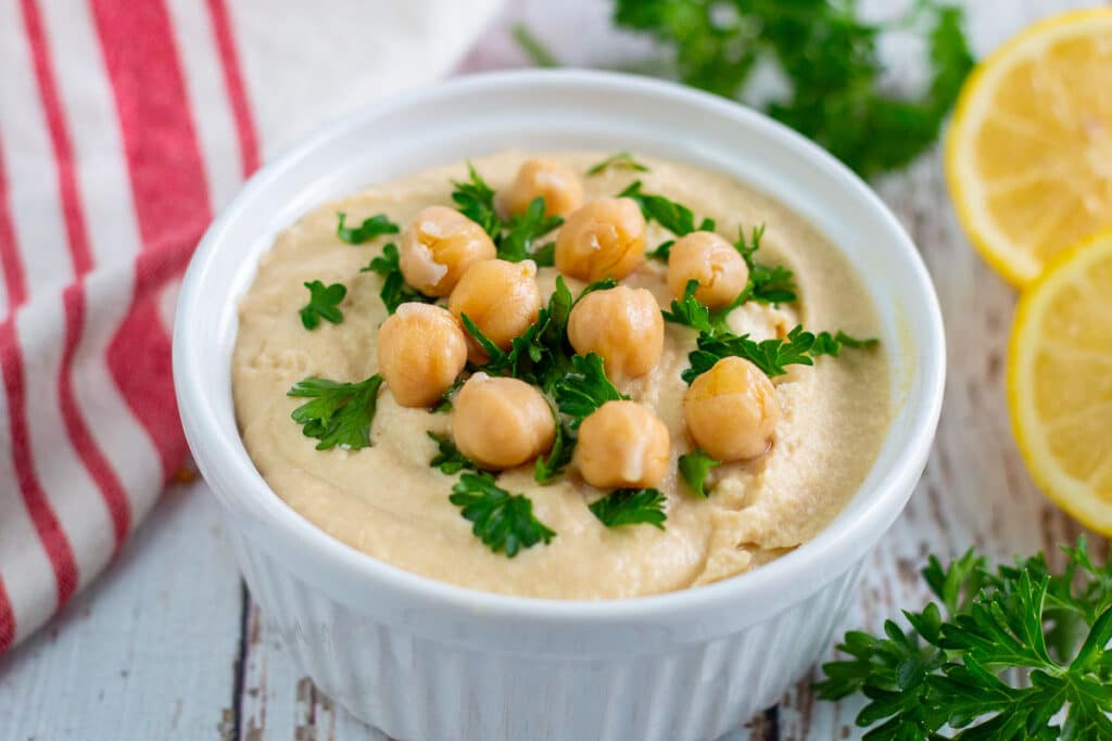 Bowl of oil free hummus garnished with chickpeas and chopped parsley surrounded by more parsley and cut lemon.