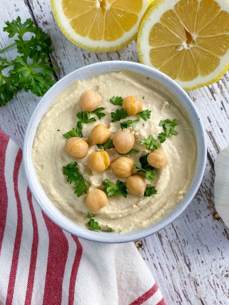 Overhead shot of bowl of hummus garnished with chickpeas and chopped parsley surrounded by more parsley and cut lemon.