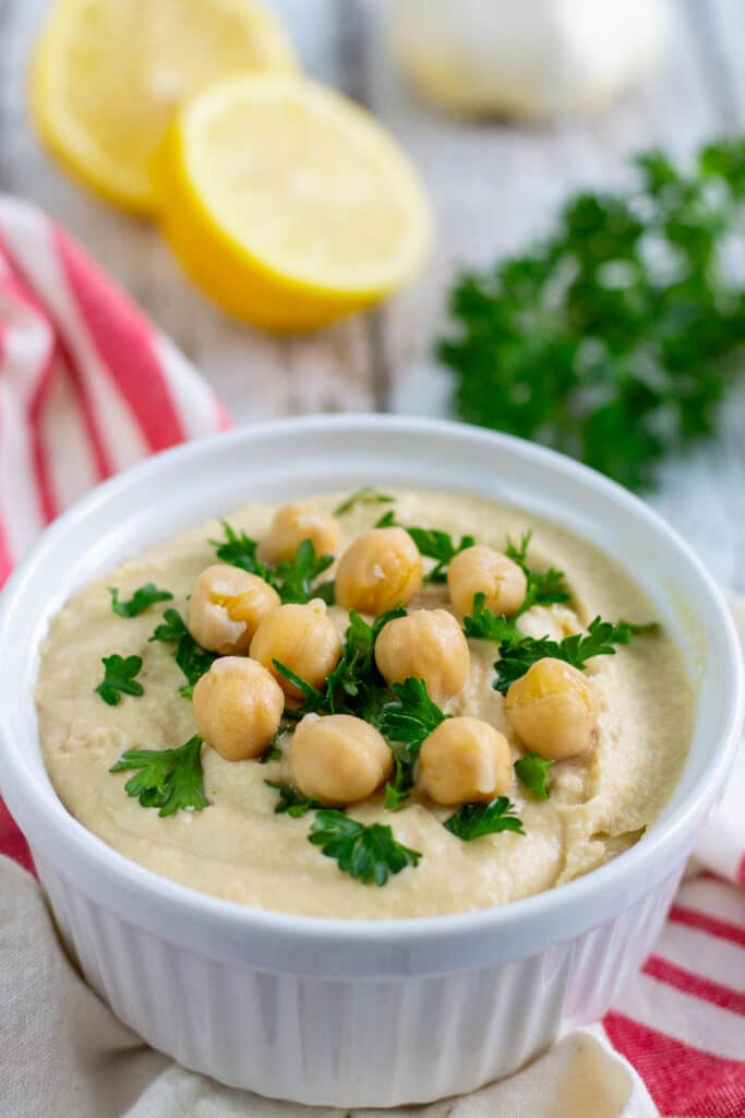 Bowl of oil free hummus garnished with chickpeas and chopped parsley with more parsley, cut lemon, and head of garlic in the background.