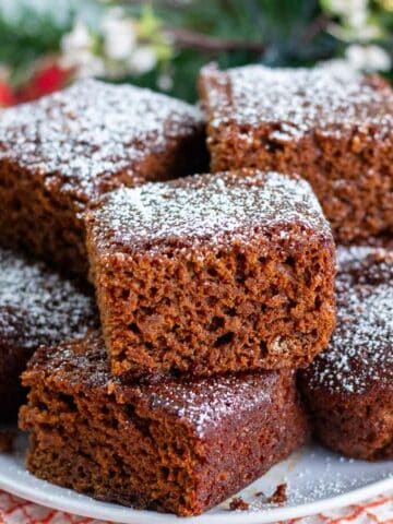 Stack of powdered sugar dusted gingerbread cake pieces o a white plate with Christmas greens in the background.