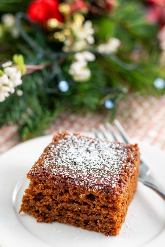 Powdered sugar dusted piece of gingerbread cake and fork on a white plate with Christmas greens in the background.