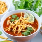 Bowl of vegan tortilla soup garnished with tortilla strips, cilantro, and lime, with bowl of tortilla strips and cilantro in the background.