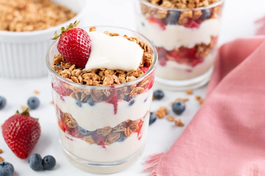 Two yogurt parfaits in front of a bowl of granola with berries and granola scattered around them.
