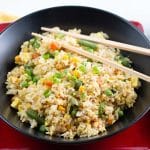 Black bowl of fried rice sitting on a square red plate with chopsticks resting in the bowl.