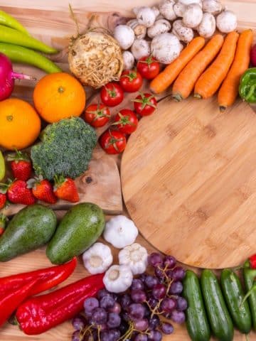 Variety of fruits and vegetables around a round wooden cutting board.