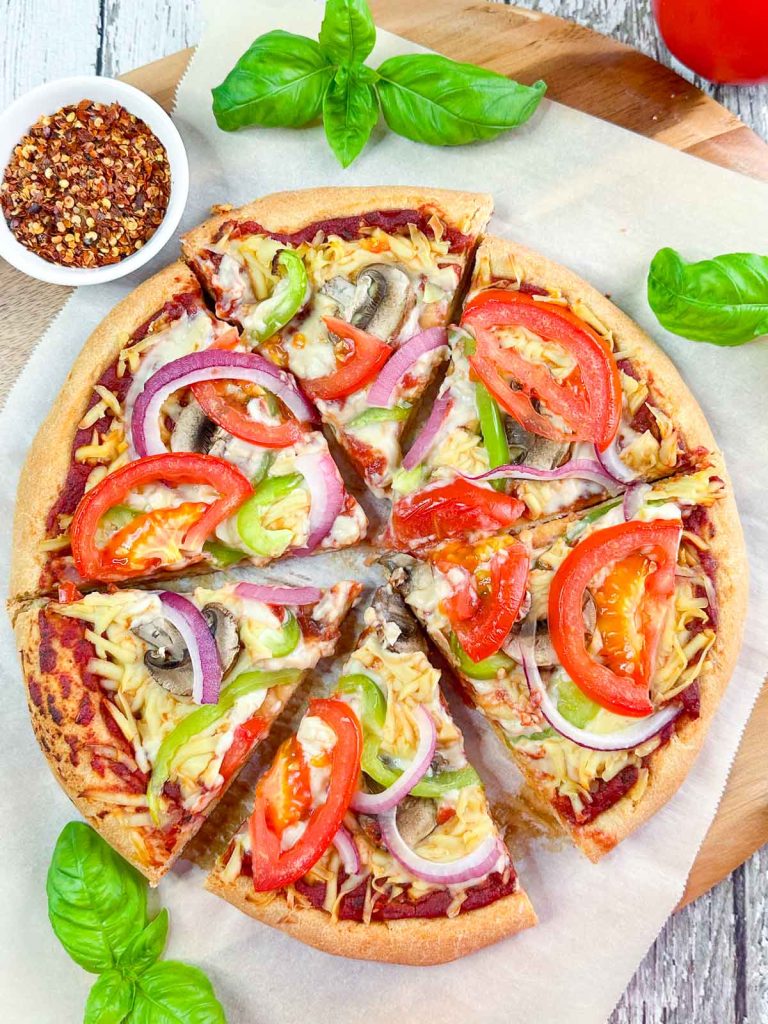 Overhead shot of sliced veggie pizza on a parchment lined pizza peel surrounded by basil and tomato.