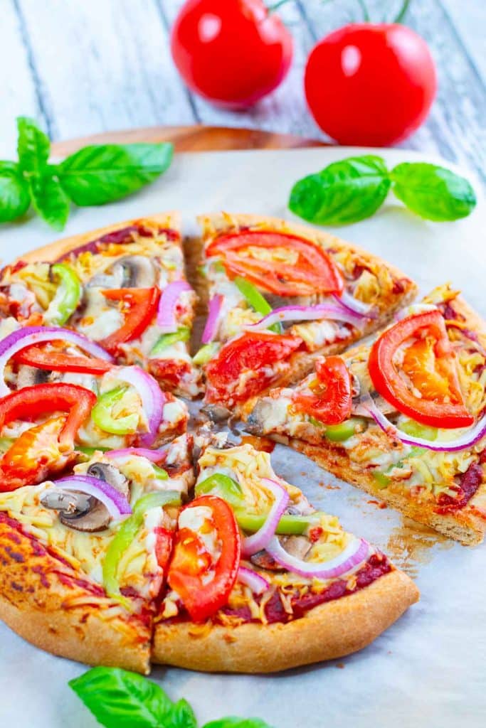 Sliced veggie pizza on a parchment lined pizza peel with basil and tomatoes in the background