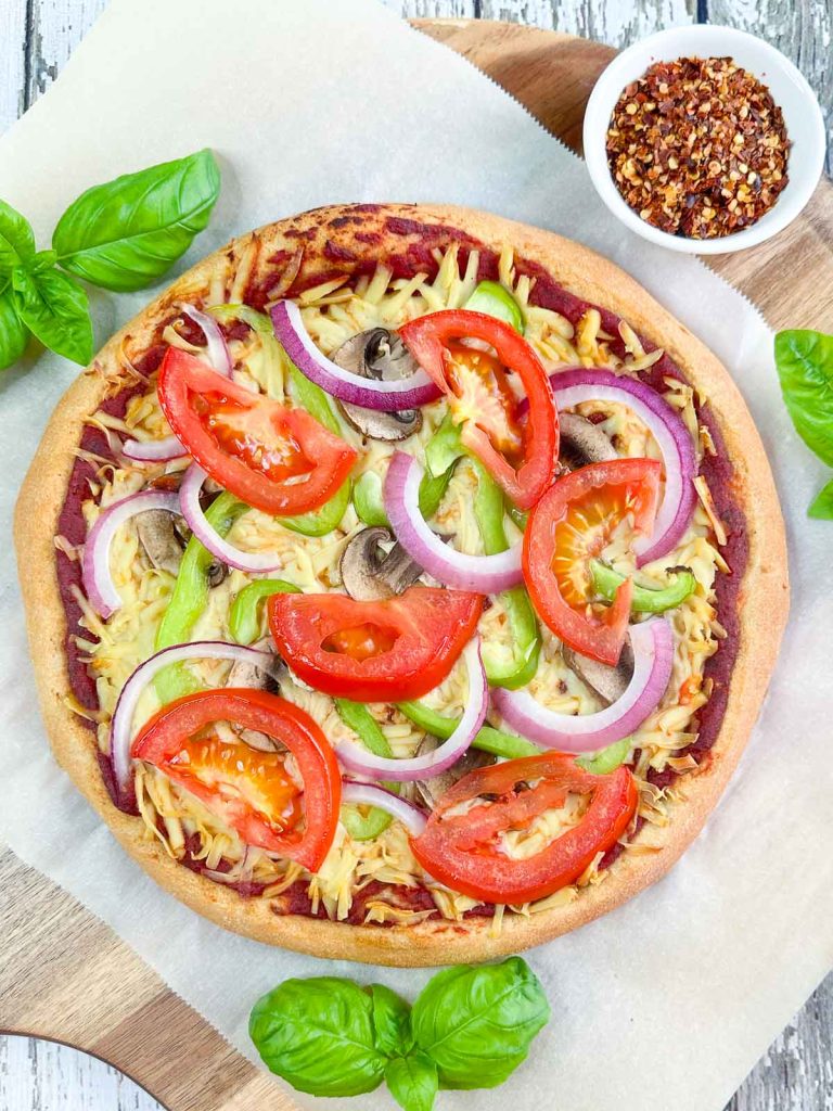 Overhead shot of unsliced vegan veggie pizza on a parchment lined pizza peel with sprigs of basil and a dish of red pepper flakes next to it.